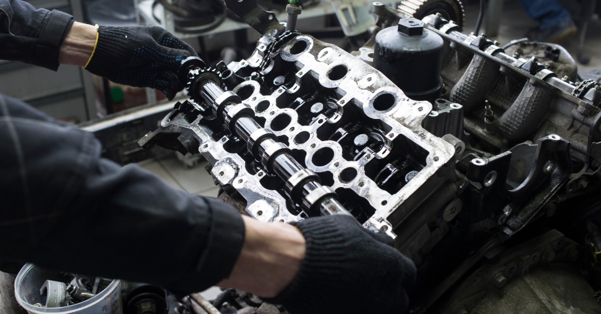A person is working on a V8 automotive engine. The engine is silver and gray, and the person wears work gloves.